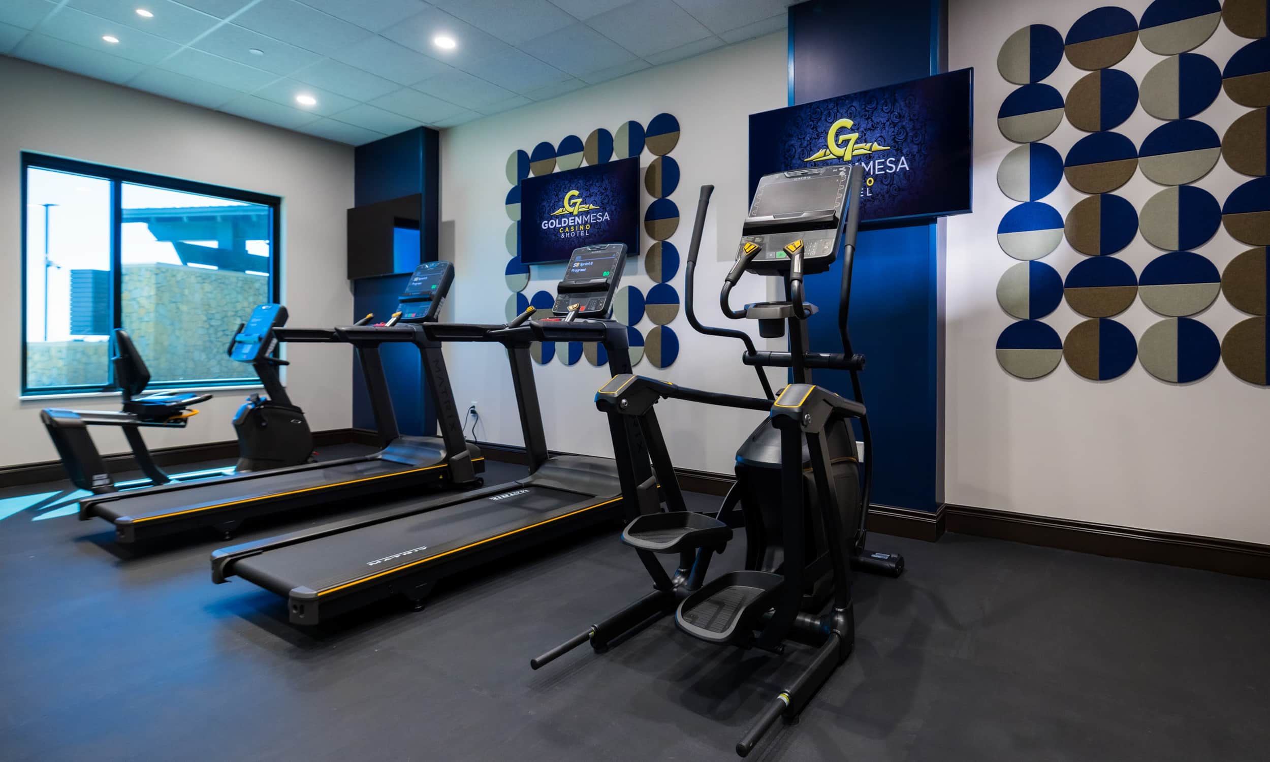 Golden Mesa Hotel fitness center with treadmills, elliptical machine, wall-mounted TVs and natural light.