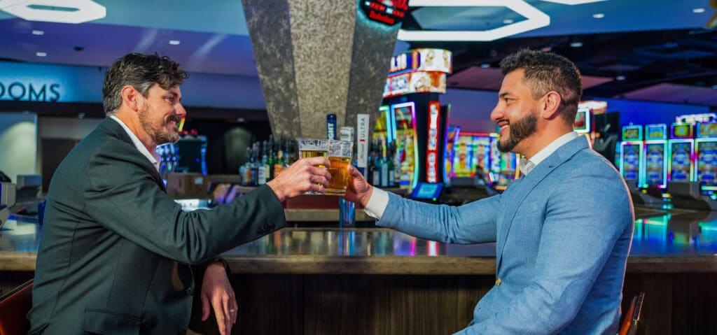 Two guests clink beers at Gold Spur Bar on the Golden Mesa Casino gaming floor with colorful slot machines in the background.