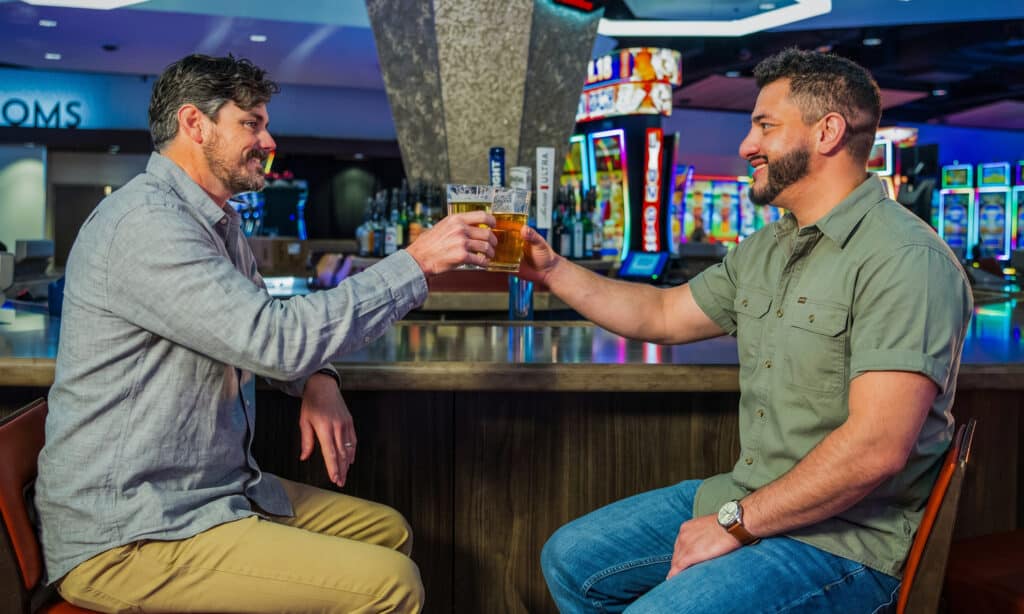 Two guests clink beers at Gold Spur Bar on the Golden Mesa Casino gaming floor with colorful slot machines in the background.