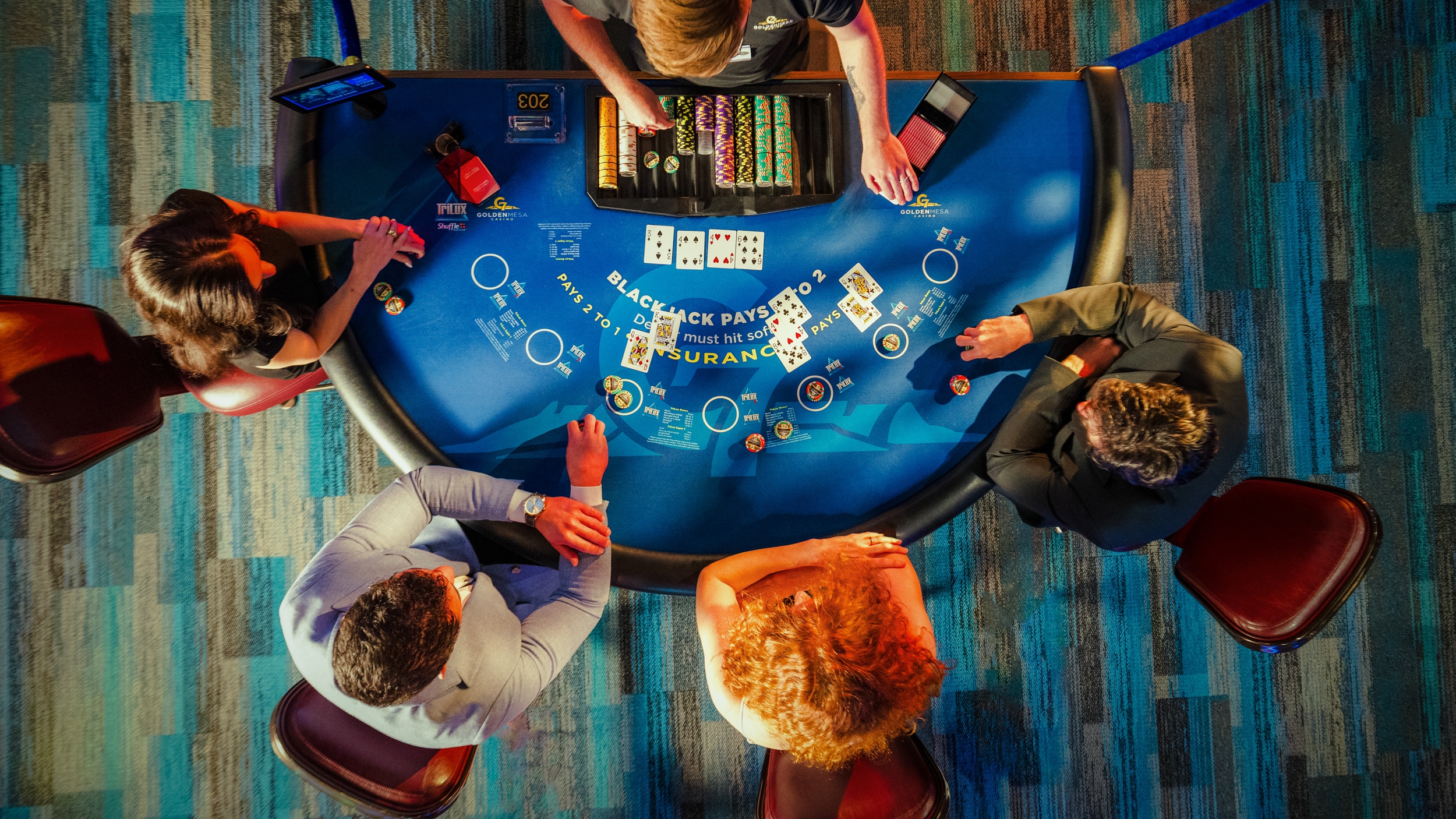 Overhead view of guests playing blackjack at a Golden Mesa Casino table with chips, cards and dealer on the casino floor.