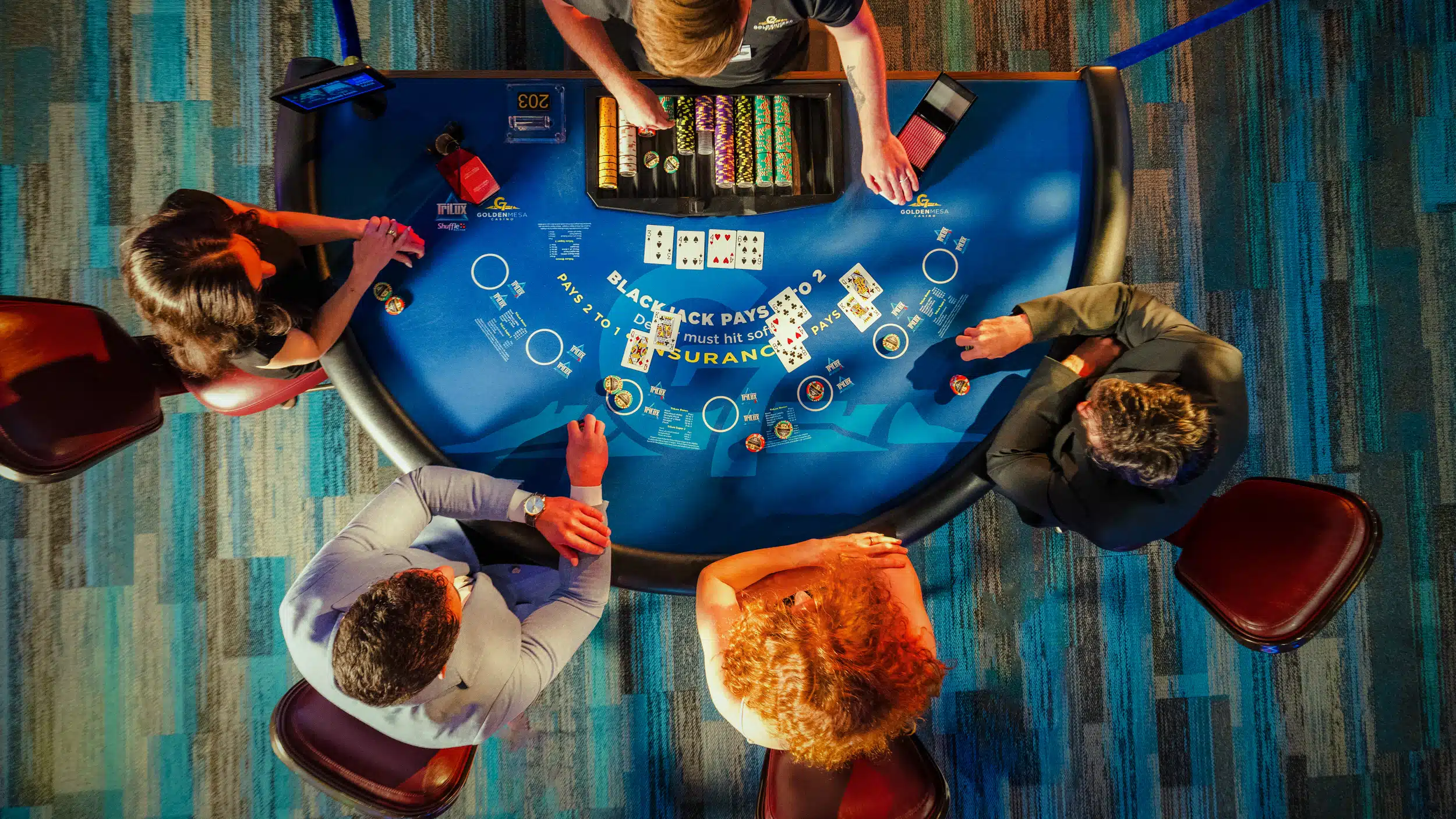 Overhead view of guests playing blackjack at a Golden Mesa Casino table with chips, cards and dealer on the casino floor.